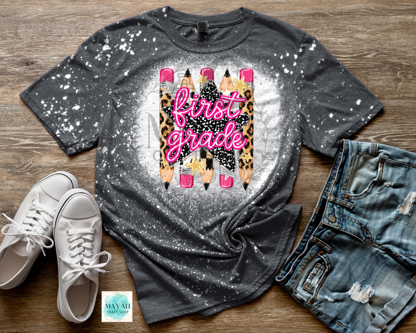 A dark gray splatter-print graphic t-shirt featuring a design of four pencils with leopard print and pink details, with the text 'first grade' in pink script. The t-shirt is styled in a flat lay on a wooden surface alongside white sneakers, distressed denim shorts.