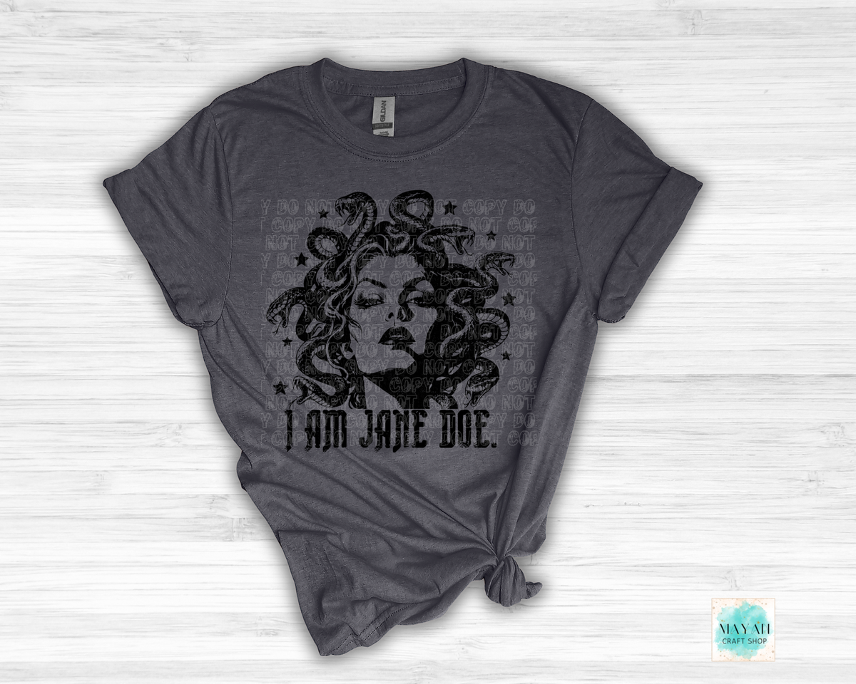 A heather charcoal t-shirt with a distressed black graphic of Medusa and the text 'I AM JANE DOE.' printed on the front, styled flat on a white wood background.