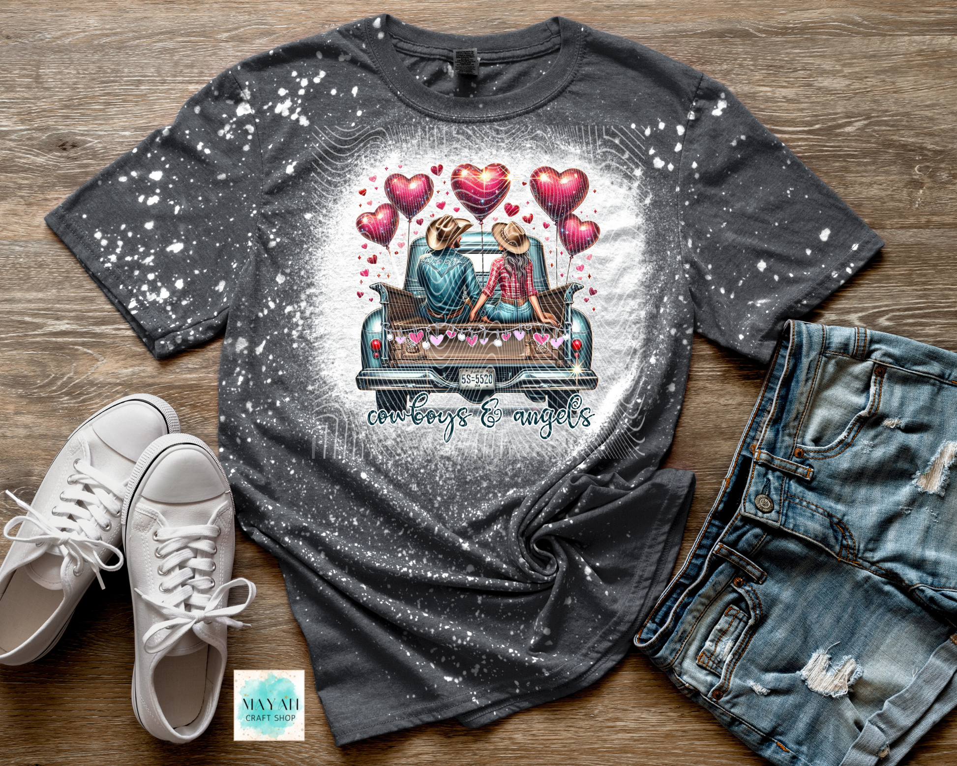 Gray t-shirt with a graphic of two people in a car surrounded by hearts, paired with white sneakers and jeans on a wooden surface.