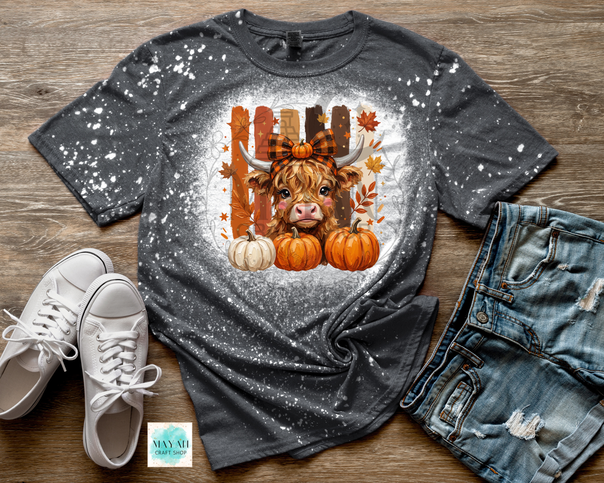 A dark gray splatter-dyed graphic t-shirt featuring a cute Highland cow with a plaid bow and autumn pumpkins, styled in a flat lay with white sneakers, distressed denim shorts.