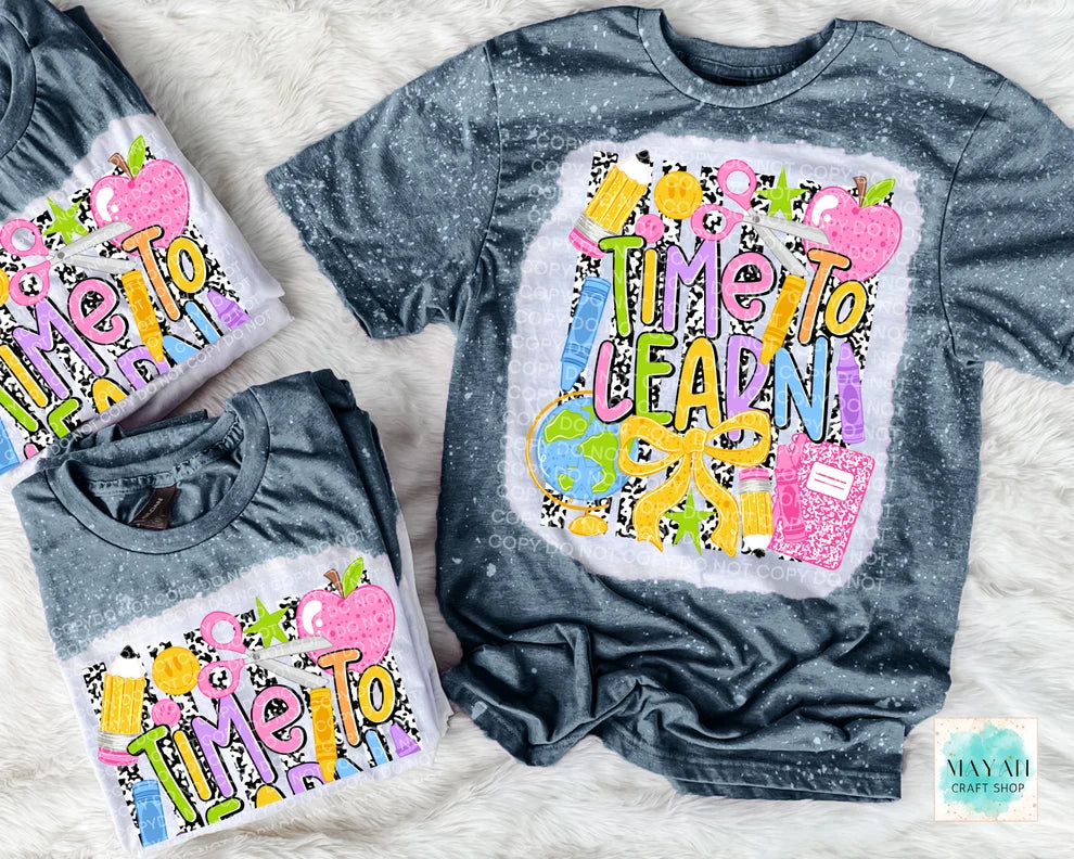 Three dark gray acid-wash t-shirts with a colorful 'Time To LEARN' graphic, arranged on a white faux fur background. The shirts feature a splattered acid-wash finish and a vibrant graphic with apples, books, and a globe.