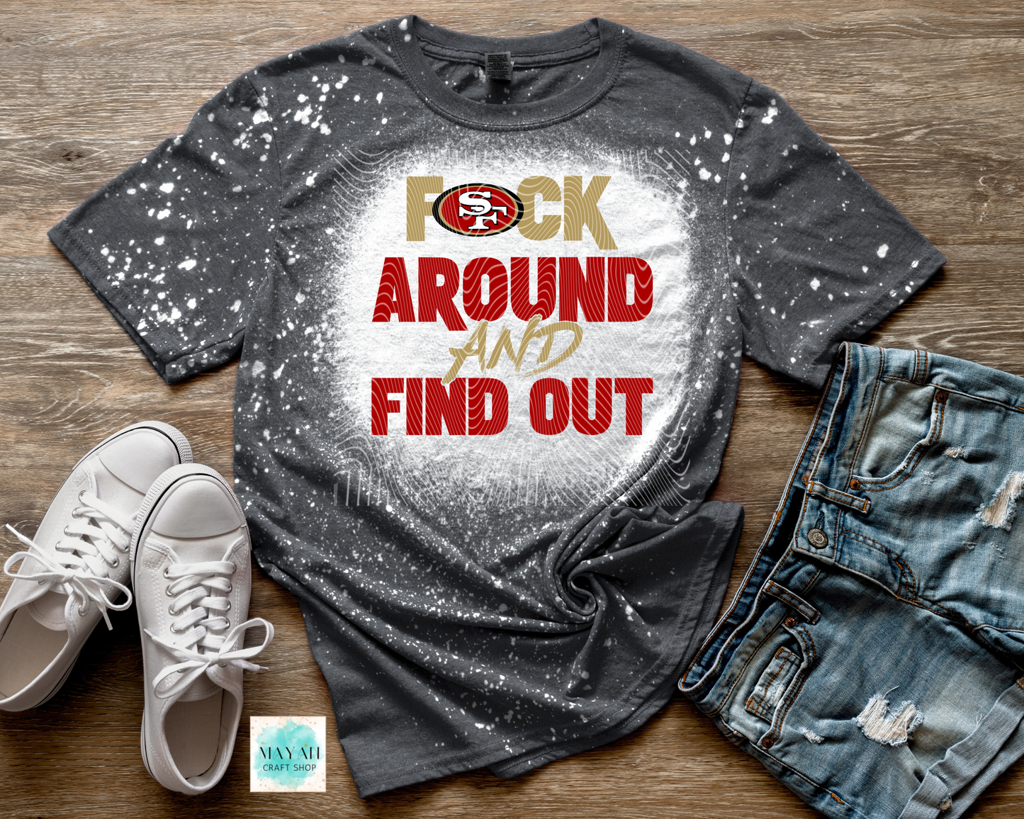 Gray bleached t-shirt with red and gold text on a wooden surface with white sneakers and blue jeans.