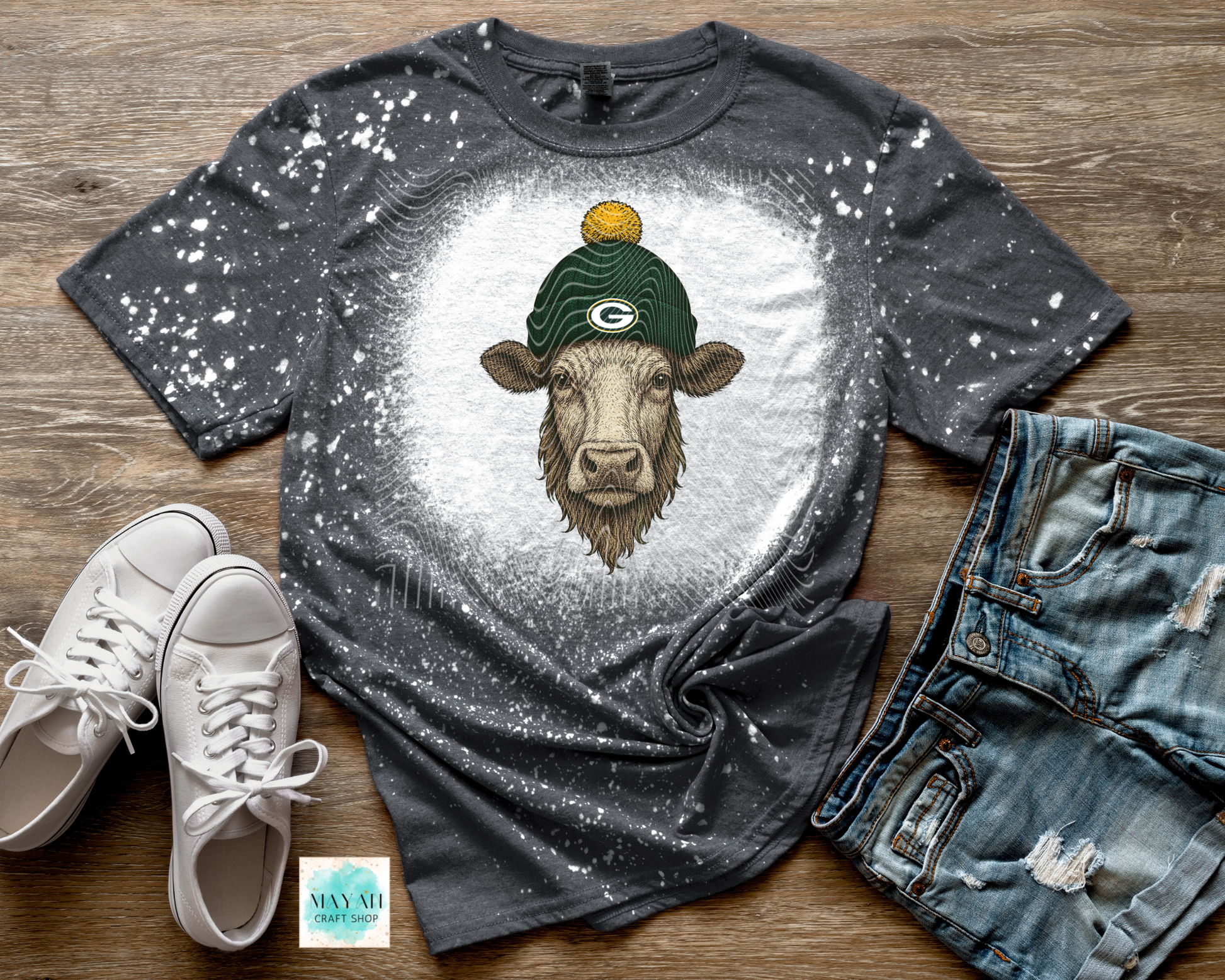 Gray t-shirt with a cow wearing a green  hat, paired with white sneakers and jeans on a wooden surface.