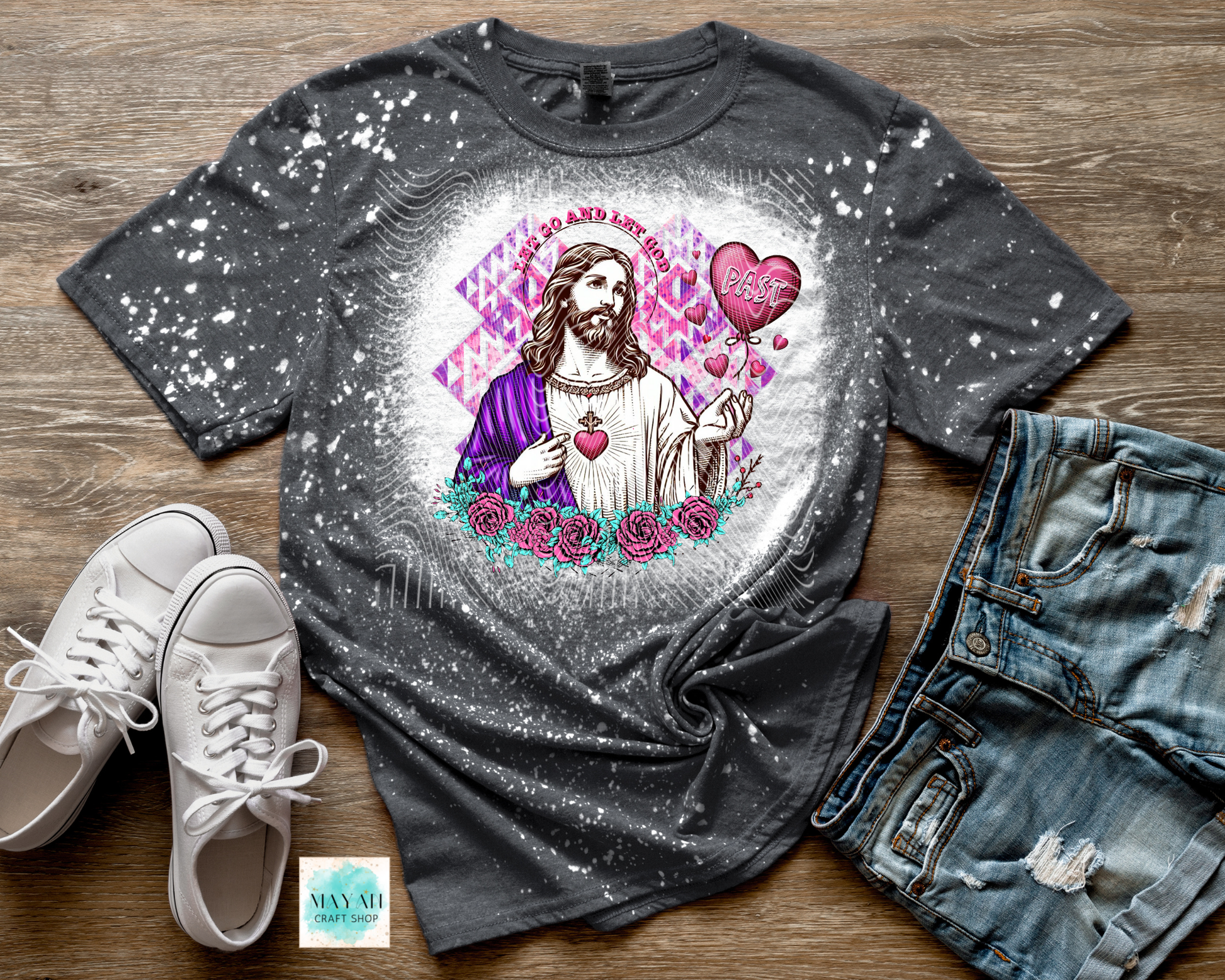 Gray t-shirt with a graphic design of a religious figure, surrounded by hearts and flowers, on a wooden surface with white sneakers and jeans.