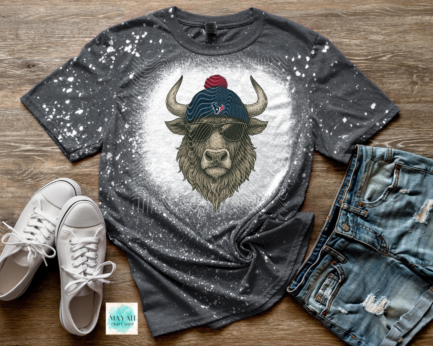 Gray t-shirt with a graphic of a bull wearing a cap and sunglasses, paired with white sneakers and jeans on a wooden surface.