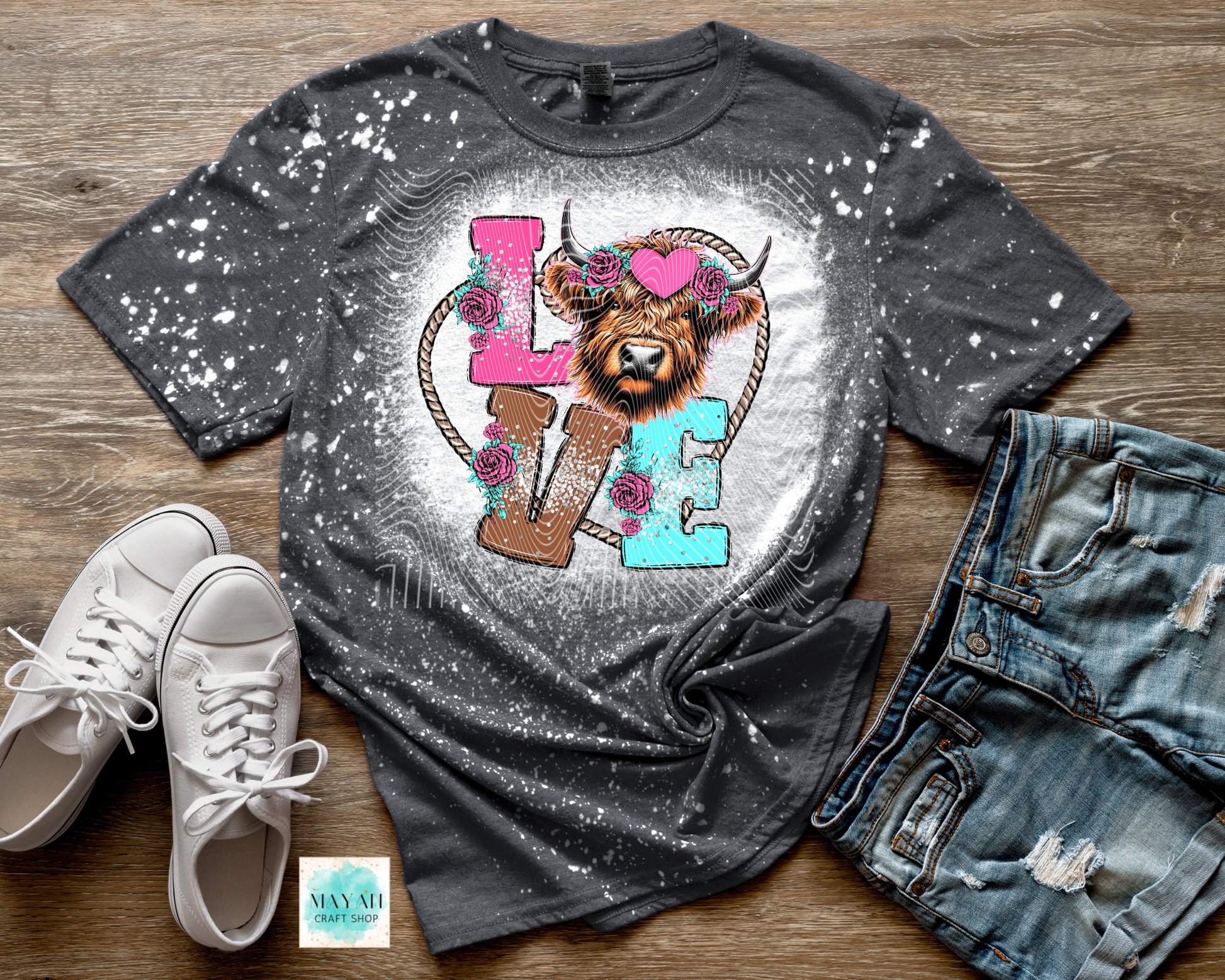 Gray t-shirt with a colorful design featuring a highland cow and flowers, paired with white sneakers and denim shorts on a wooden surface.