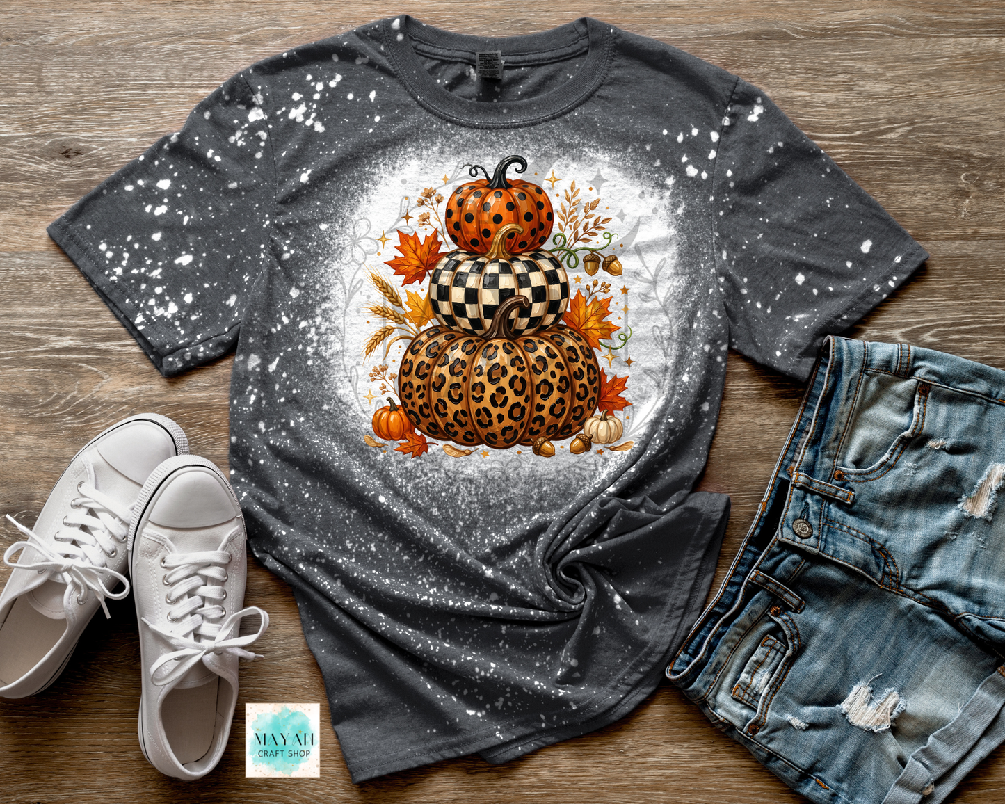 A dark gray splatter-print t-shirt featuring a stacked autumn pumpkin graphic with leopard print, checkerboard pattern, and orange leaves. The shirt is styled in a flat lay on a wooden background alongside white sneakers, distressed denim shorts.