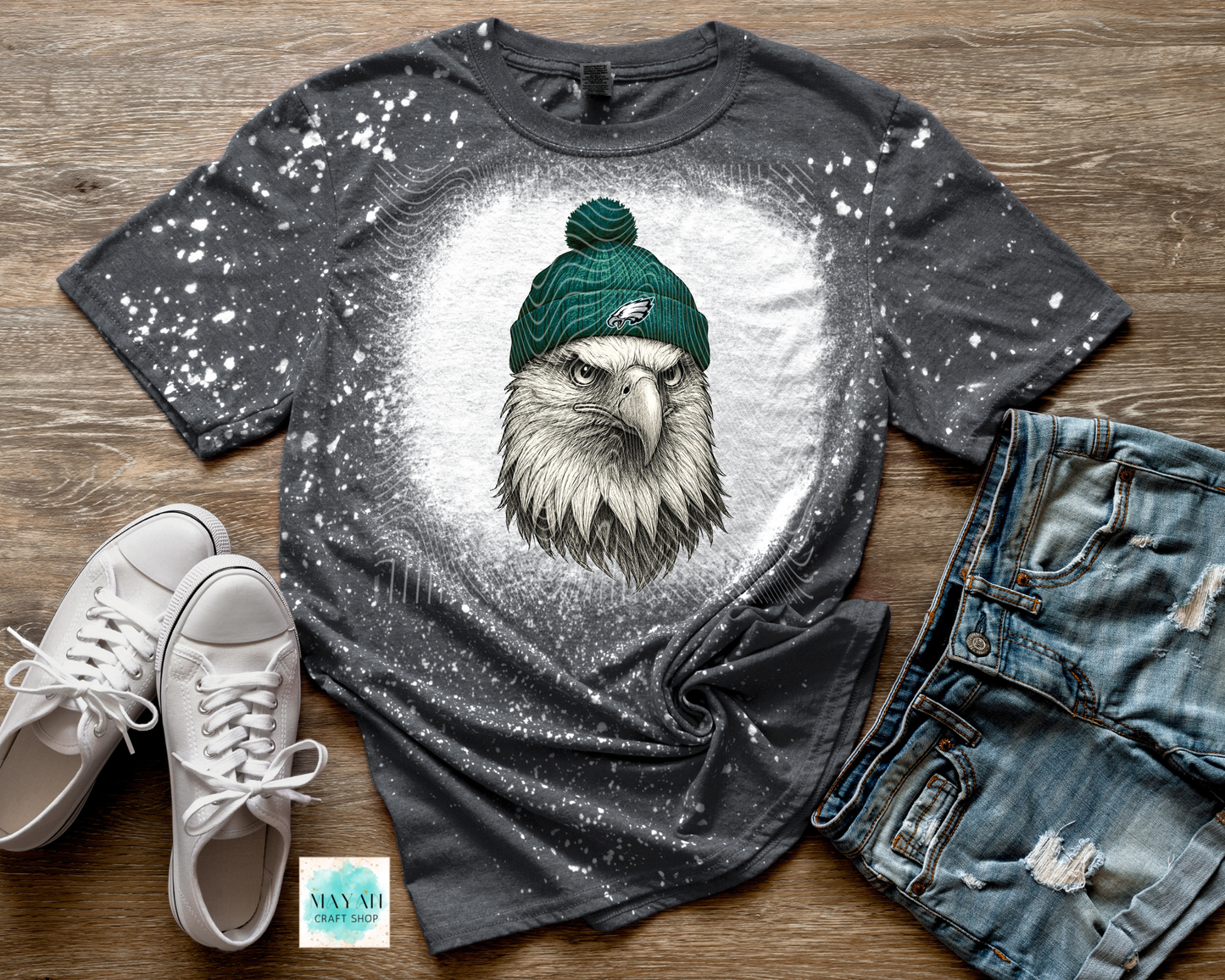 Gray t-shirt with an eagle wearing a green beanie, paired with white sneakers and jeans on a wooden surface.