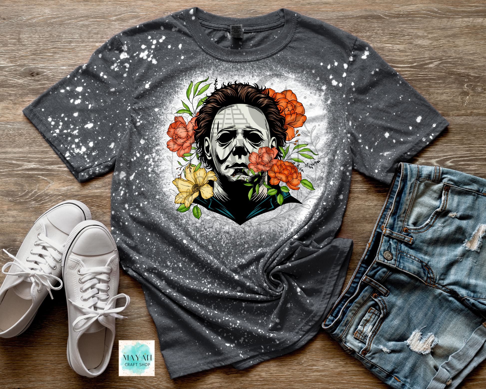 Gray t-shirt with a graphic design of a face surrounded by flowers, paired with white sneakers and denim shorts on a wooden surface.