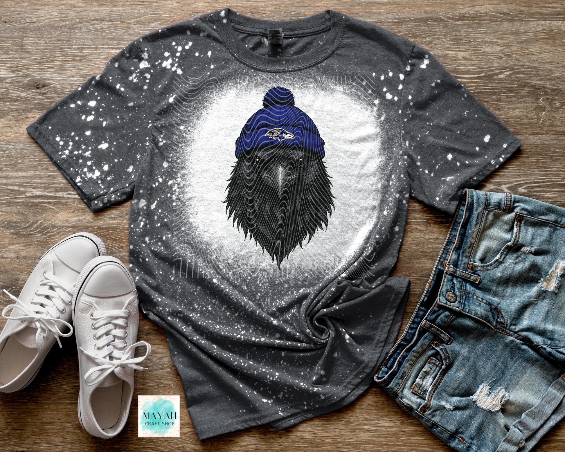Gray t-shirt with a black crow wearing a blue beanie on a wooden surface with white sneakers and jeans.