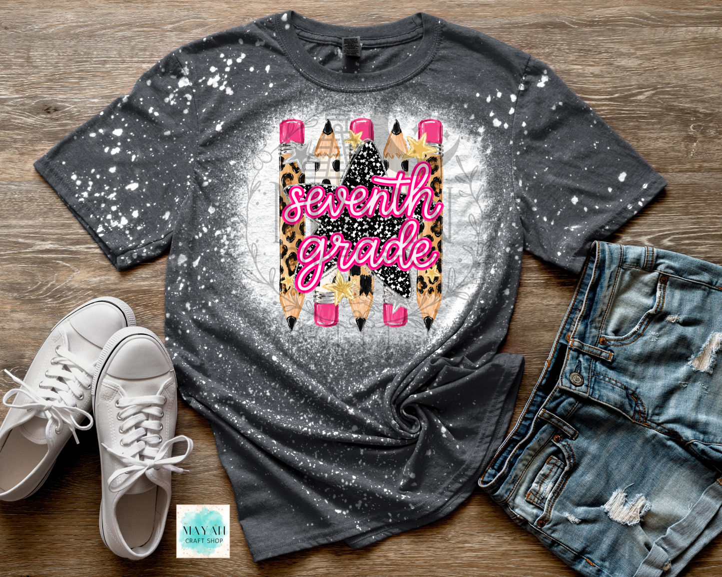 A dark gray splatter-print graphic T-shirt laid flat on a wood surface, styled with white sneakers, distressed denim shorts. The shirt features a central graphic of colorful pencils with leopard print and the words 'seventh grade' in pink script.