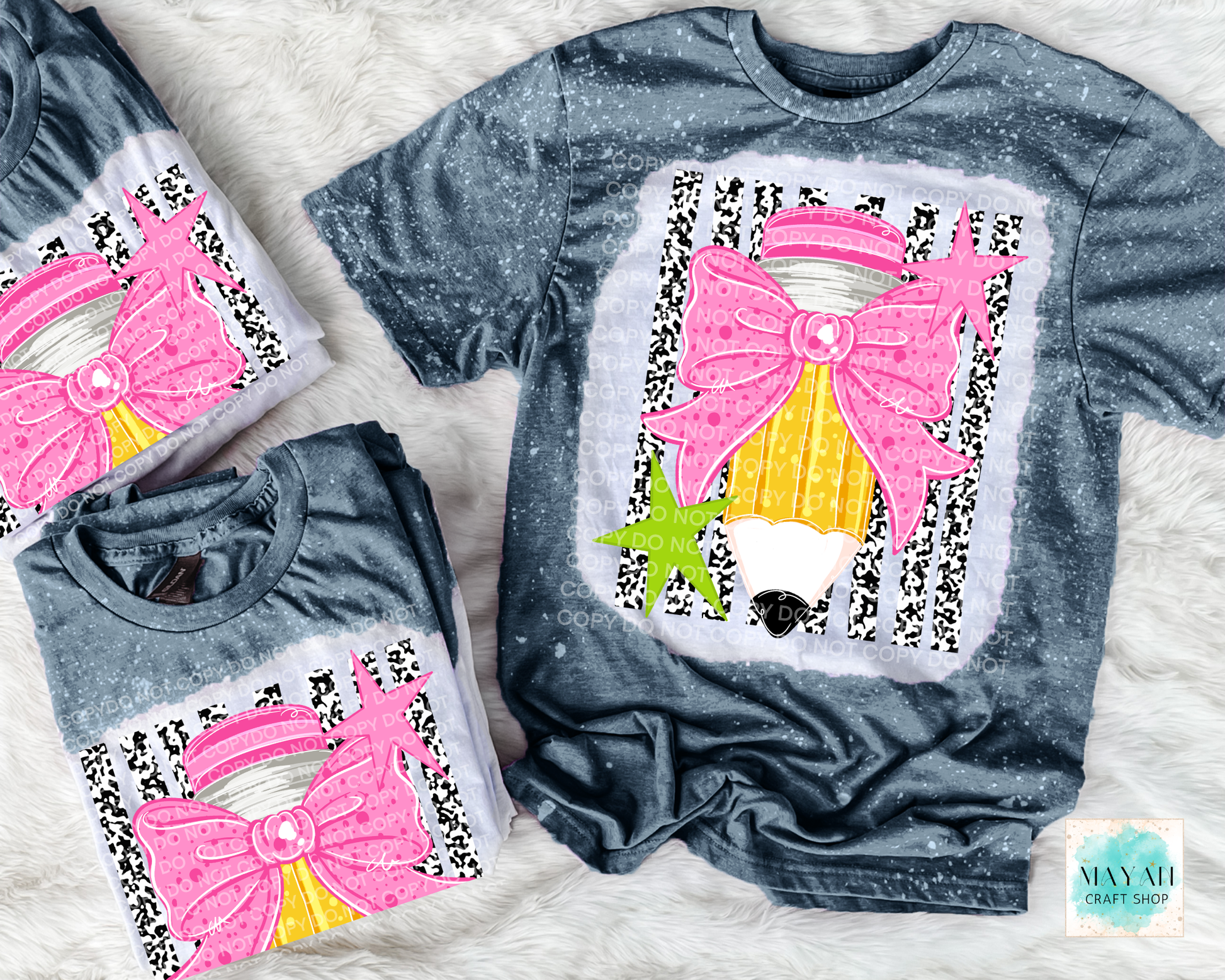 Three gray short-sleeve graphic T-shirts arranged on a white faux-fur surface. The shirts feature a large applique design of a pink bow, pencil, and green star on a black-and-white patterned background.