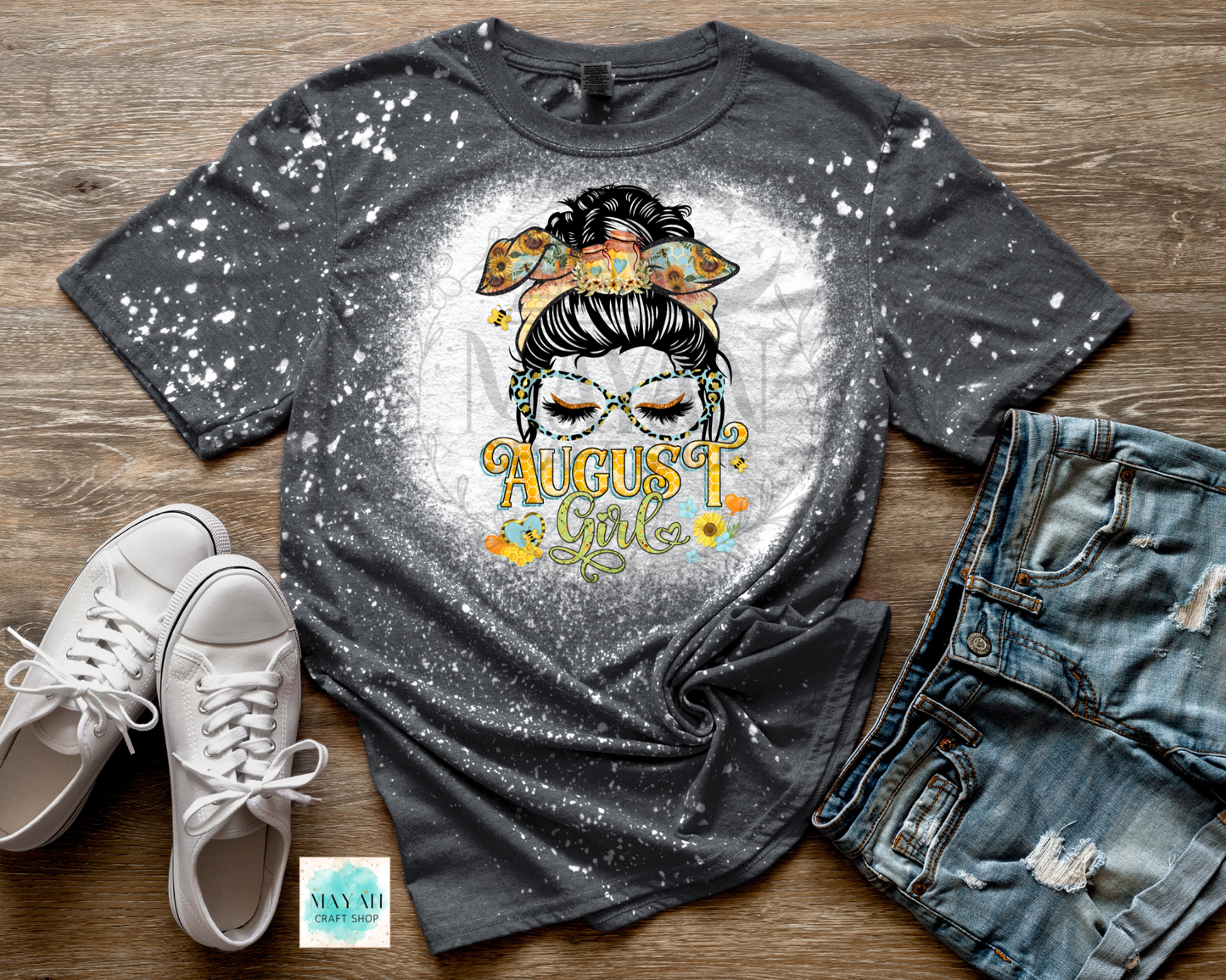 A dark gray splatter-print graphic t-shirt featuring a sugar skull illustration and the text 'August Girl' in yellow, styled in a flat lay with white sneakers, distressed denim shorts.