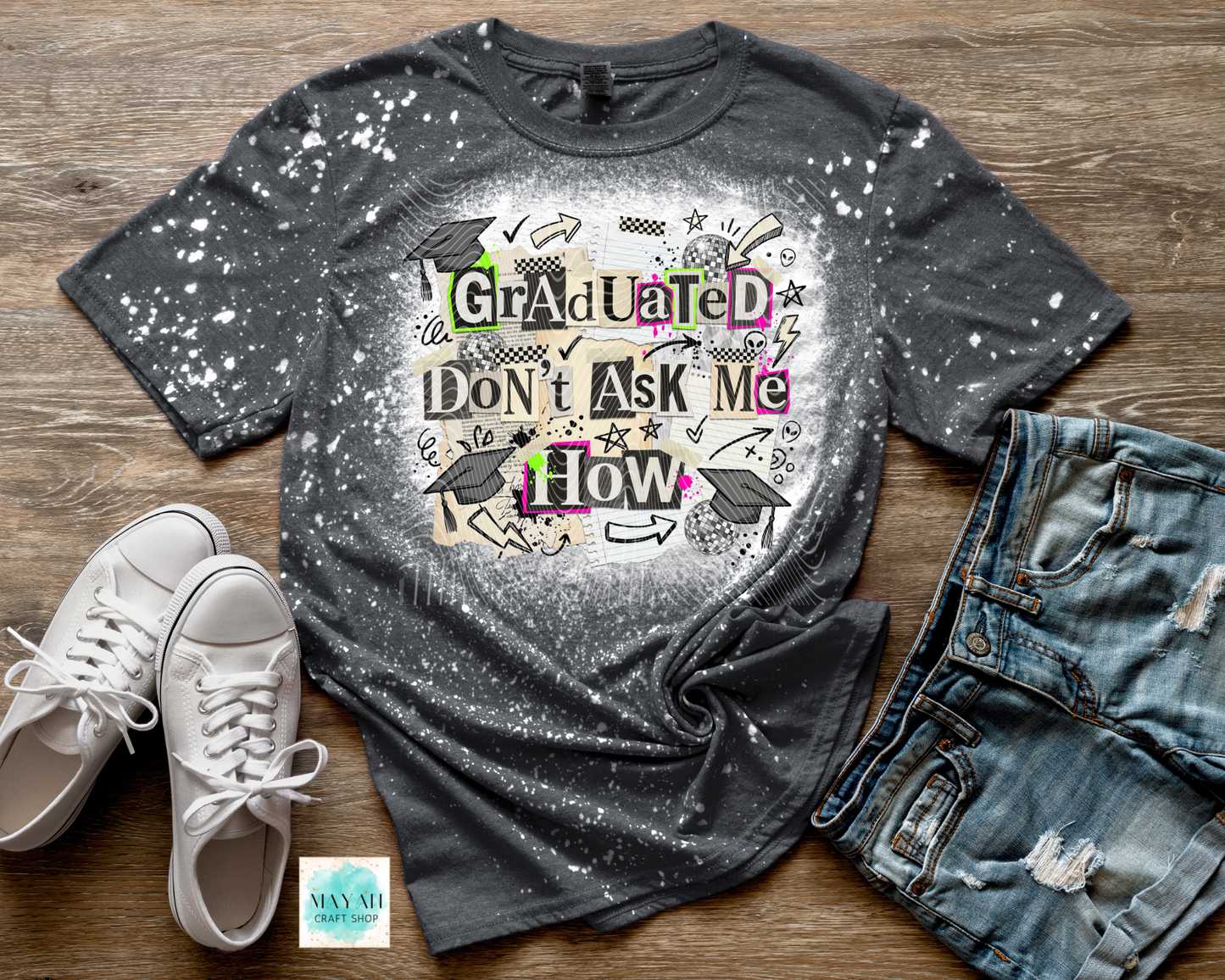Gray t-shirt with graduation-themed design on a wooden surface with shoes and jeans.
