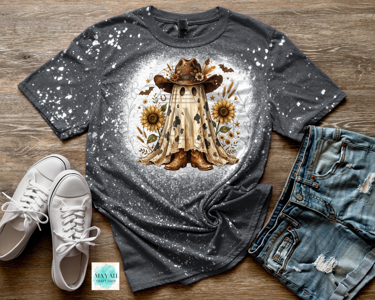 A dark gray splatter-print t-shirt with a central graphic of a cowboy in a poncho surrounded by sunflowers, styled in a flat lay with white sneakers, distressed denim shorts.