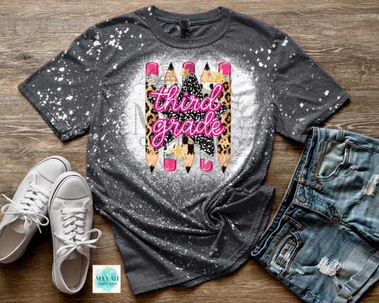 A dark gray splatter-print graphic t-shirt featuring a design of four pencils with leopard print and pink details, with the text 'third grade' in pink script. The t-shirt is styled in a flat lay on a wooden surface alongside white sneakers, distressed denim shorts.