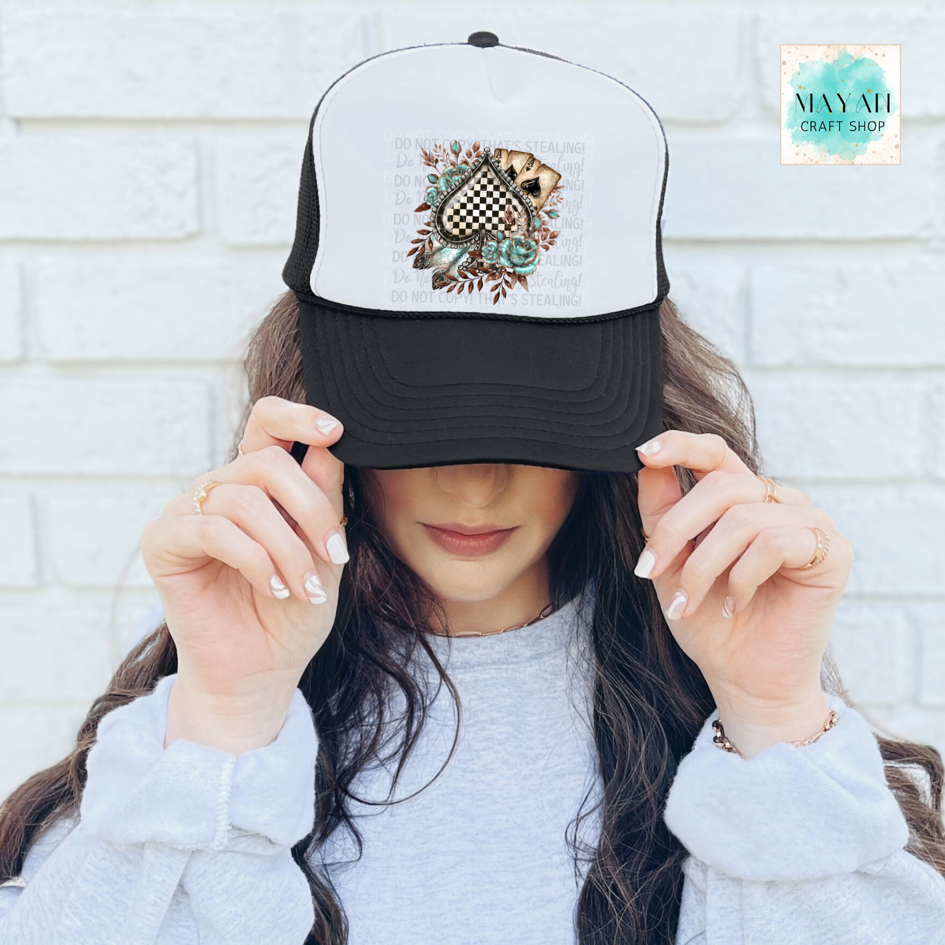 Person wearing a cap with a design, holding it up to their face against a white brick wall.