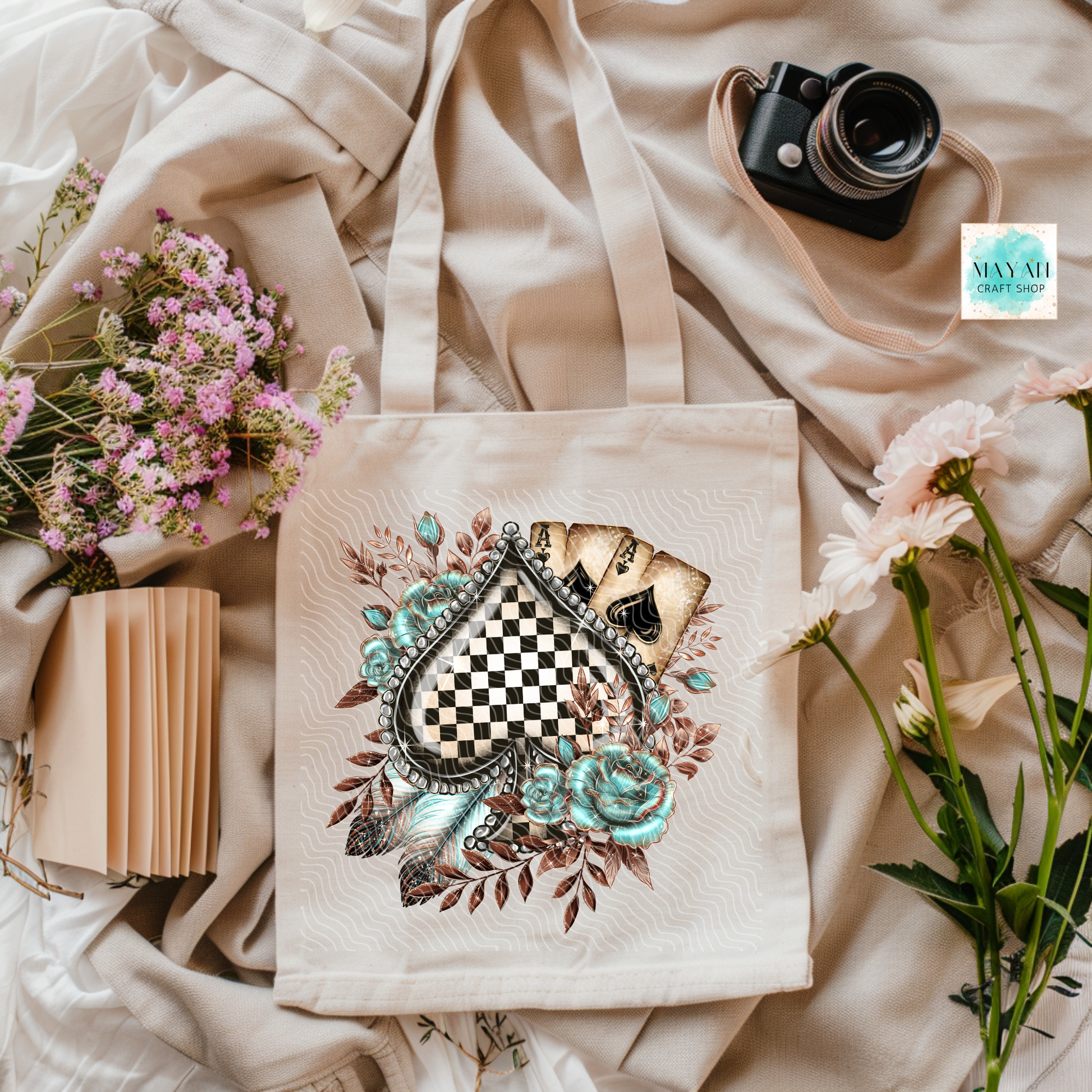 Tote bag with decorative design on a soft fabric background with flowers and a camera.