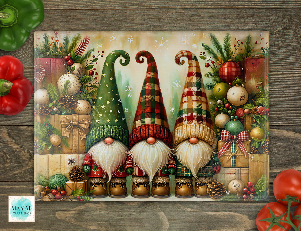 A cutting board featuring three festive gnomes with tall patterned hats, white beards, and boots, surrounded by ornaments, gifts, pinecones, and evergreen branches. The print is photographed flat on a rustic wood surface with decorative red and green peppers in the corners.