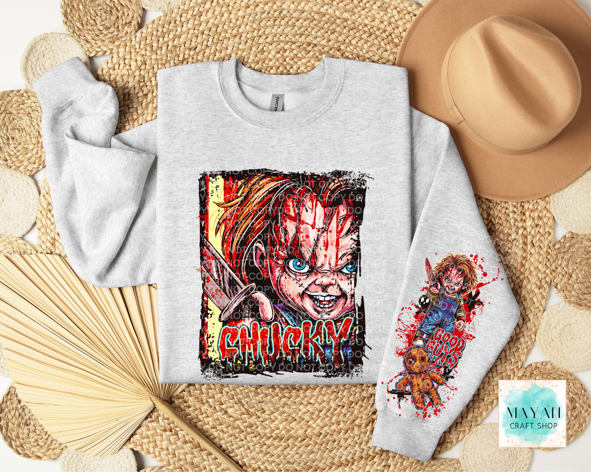 A light gray sweatshirt laid flat on a woven mat, featuring a large horror-style face graphic on the front and a smaller print on the sleeve. A tan hat, folded fan, and woven textile props surround the sweatshirt.