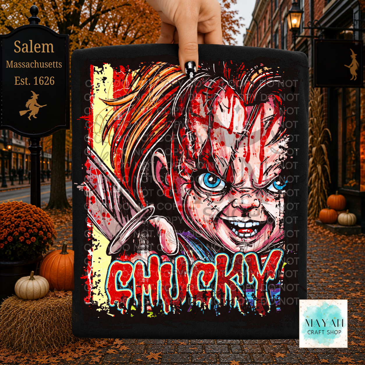 A hand holds a black shirt featuring a horror-style illustration with a knife, blue eyes, red blood effects in large dripping red letters. The background is a Halloween-themed street scene with autumn leaves, pumpkins, a sign, and storefronts.