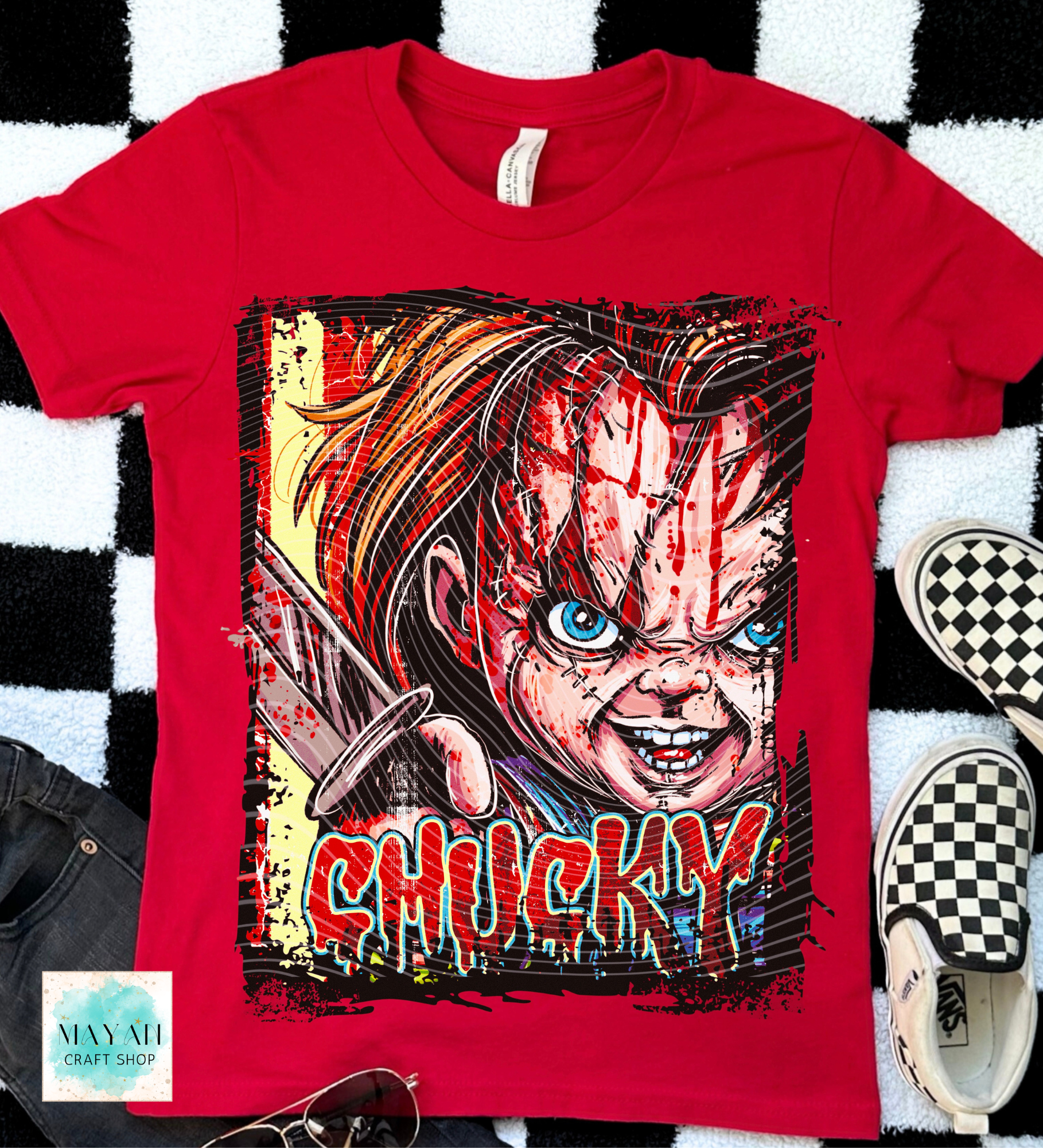 A red short-sleeve graphic T-shirt laid flat on a black-and-white checkered surface, featuring a large horror-style print in red dripping letters. 