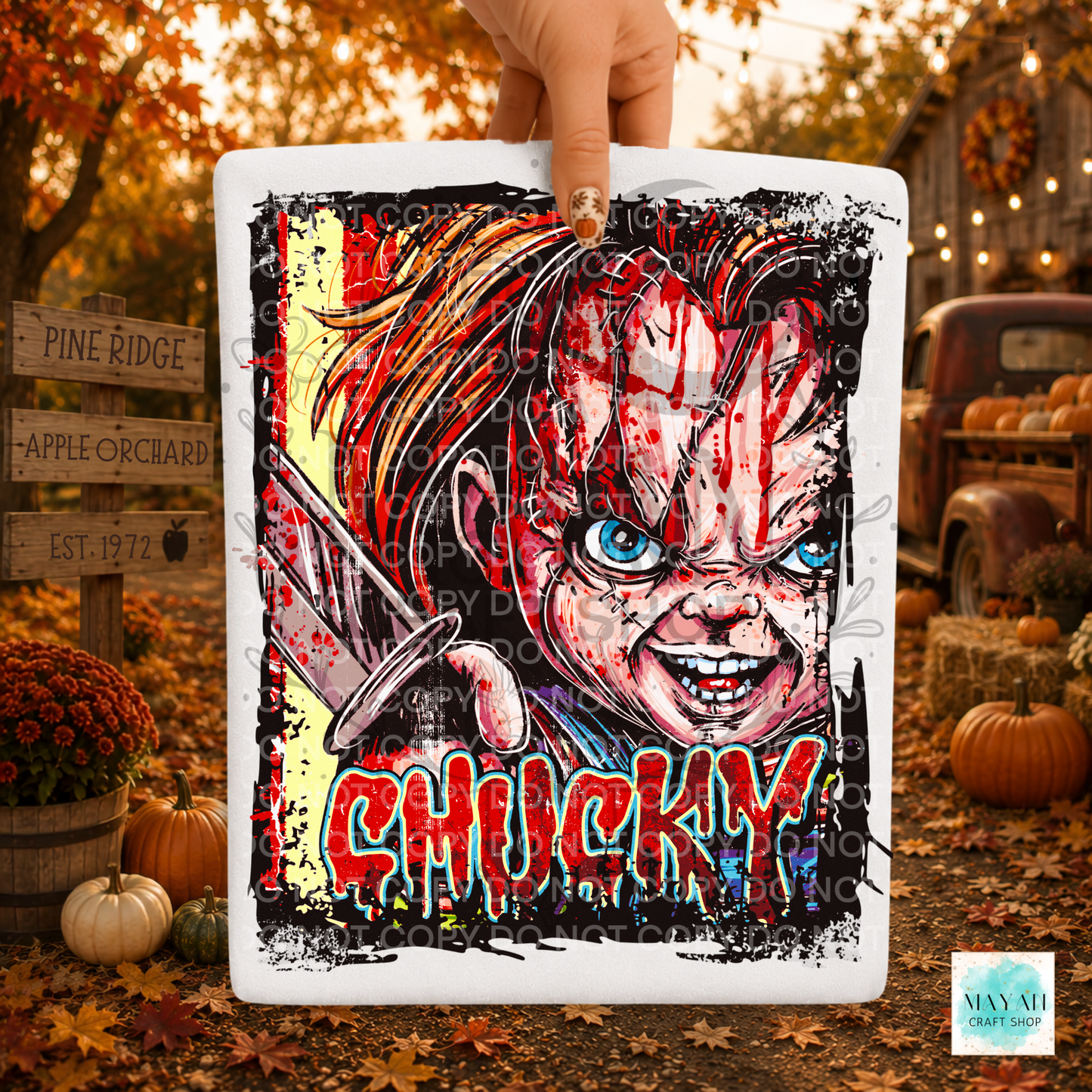 A hand holds a square white shirt with red blood splatter, blue eyes, and the in dripping red letters. The scene is set outdoors in autumn with pumpkins, fall leaves, and rustic decorations in the background.