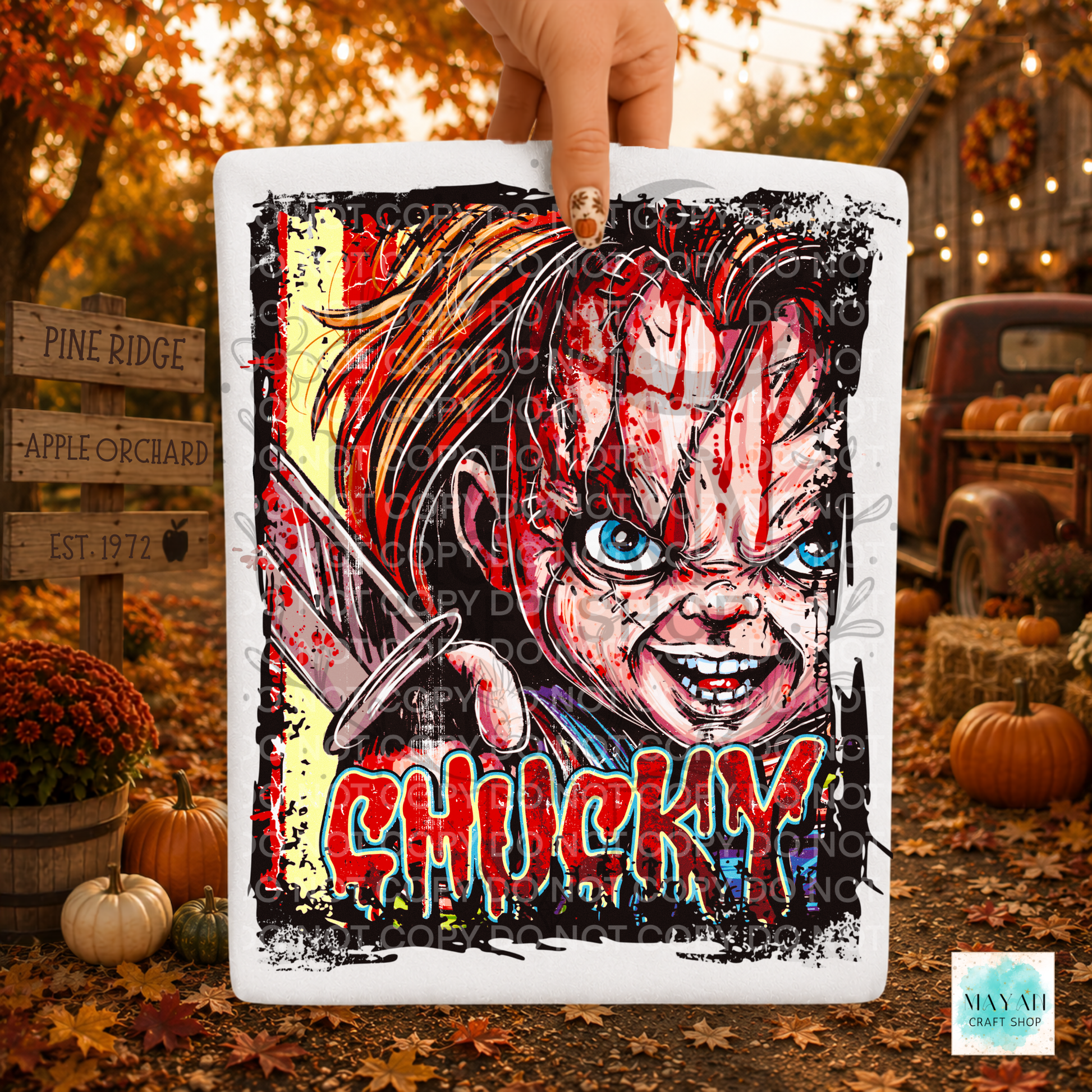 A hand holds a square white shirt with red blood splatter, blue eyes, and the in dripping red letters. The scene is set outdoors in autumn with pumpkins, fall leaves, and rustic decorations in the background.