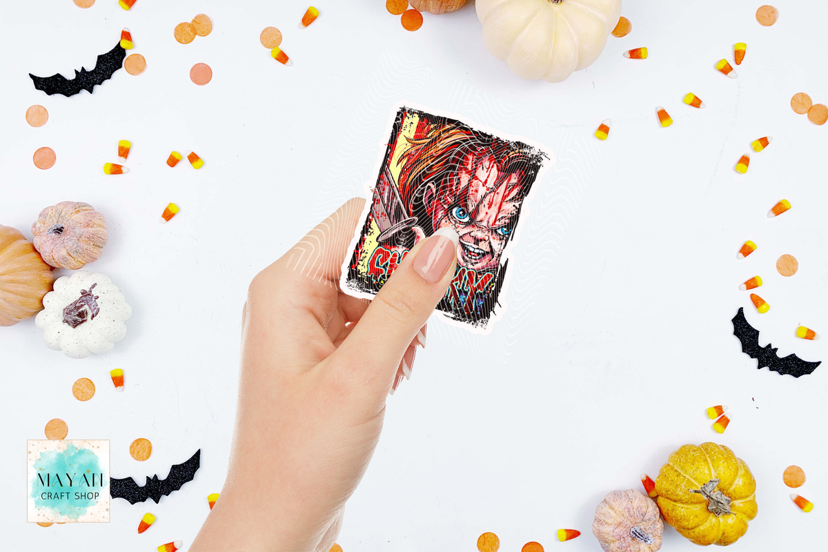 A hand holds a rectangular sticker or card featuring a horror character illustration with red hair and a black-and-white face. The background is a white flat-lay styled with Halloween props including small pumpkins, candy corn, black bat cutouts, and orange confetti.