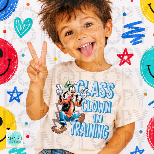 A smiling young boy with his tongue sticking out makes a peace sign while wearing a cream-colored t-shirt with a cartoon graphic and the text 'CLASS CLOWN IN TRAINING'. The background is a white patterned surface with colorful faces, stars, and scribbles, overlaid with a faint diagonal watermark.