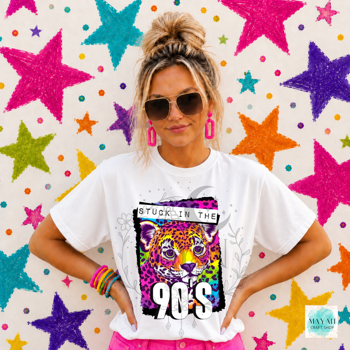 A woman wearing a white graphic t-shirt with a colorful 90s leopard print design, standing in front of a white wall decorated with colorful glittery stars.
