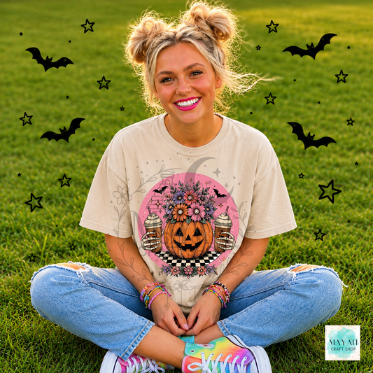 Person wearing a Halloween-themed t-shirt with a pumpkin and coffee design, sitting on grass with decorative elements.