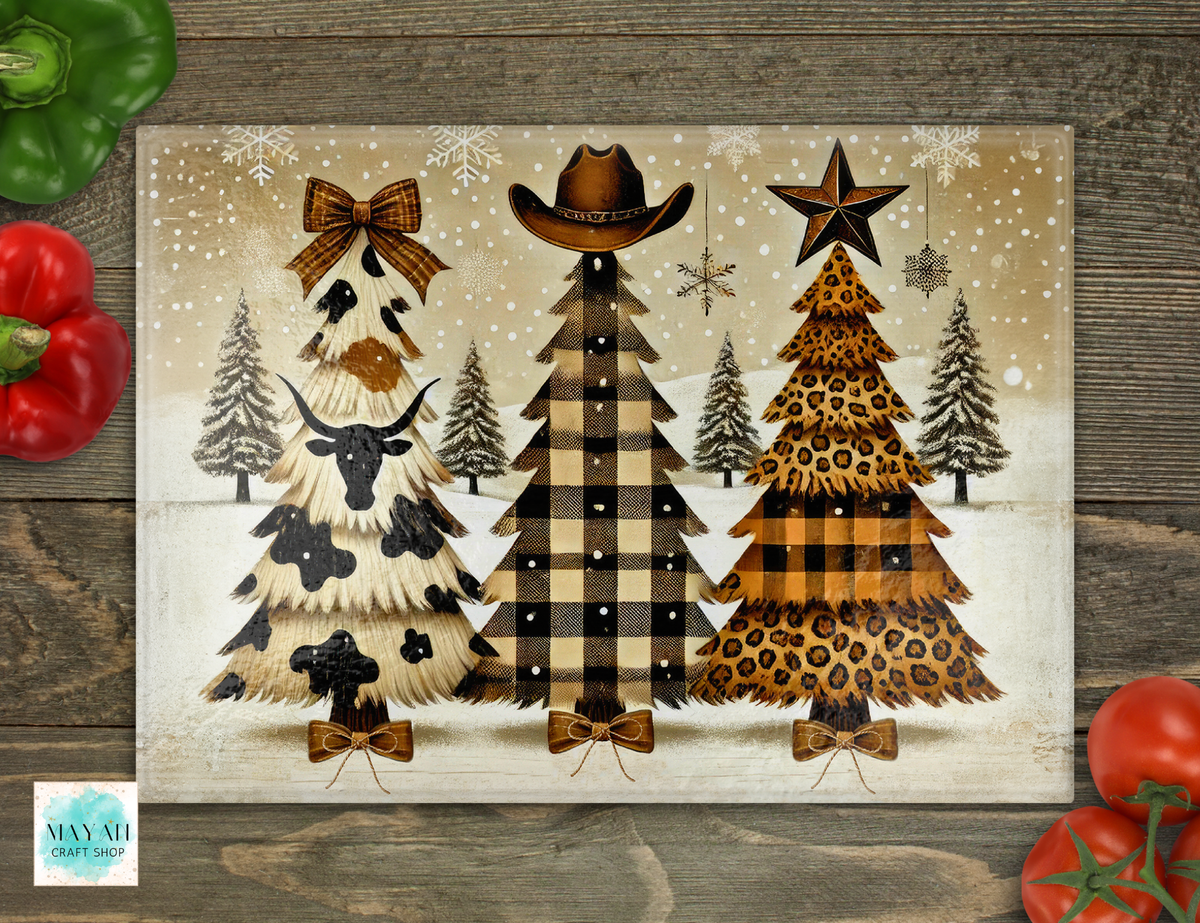A rustic Christmas-themed cutting board featuring three stylized Christmas trees in cowgirl and leopard print, set on a weathered wood background with decorative red and green peppers in the corners.