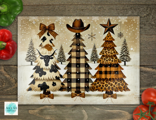 A rustic Christmas-themed cutting board featuring three stylized Christmas trees in cowgirl and leopard print, set on a weathered wood background with decorative red and green peppers in the corners.