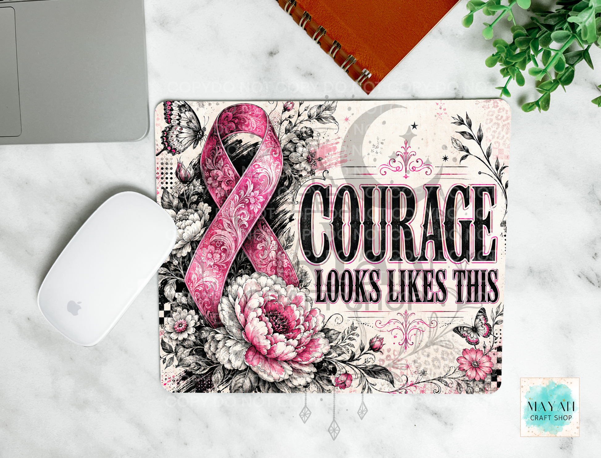 A mouse pad with a floral and ribbon design sits on a white marble surface. The mat features the words 'COURAGE LOOKS LIKES THIS' alongside a pink paisley awareness ribbon, black-and-white flowers, butterflies, and a crescent moon.