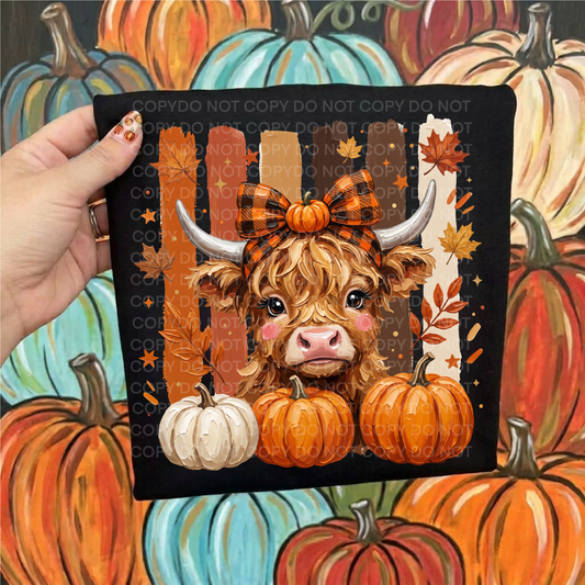 A hand holds a black shirt, featuring a cartoon Highland cow with orange plaid hair bow, pumpkins, and autumn leaves. The background is a colorful painted pattern of pumpkins.