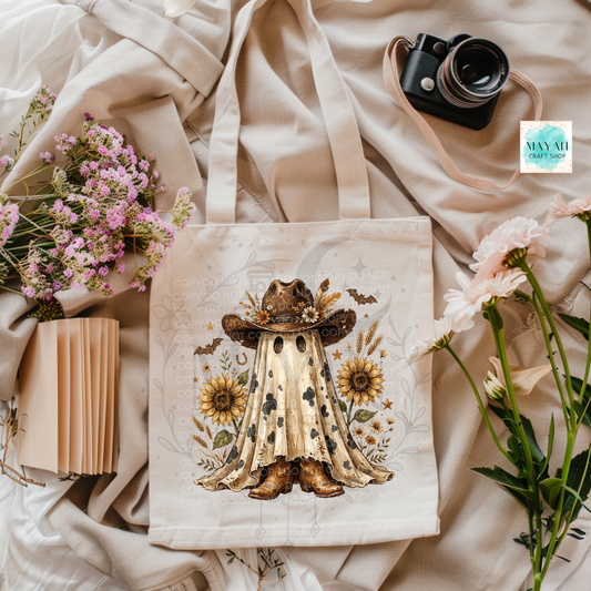A cream canvas tote bag with a western ghost illustration, styled in a flat lay with flowers, a camera, and a notebook on a beige fabric background.