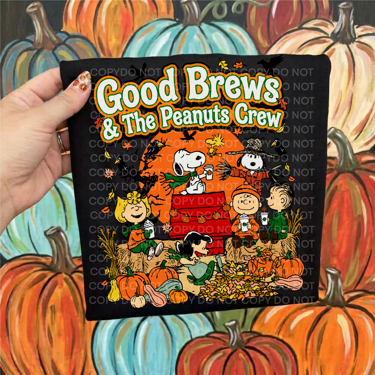A hand holds a black shirt featuring a colorful Fall graphic with the text 'Good Brews' over a pumpkin illustration. The background is a painted scene of pumpkins.