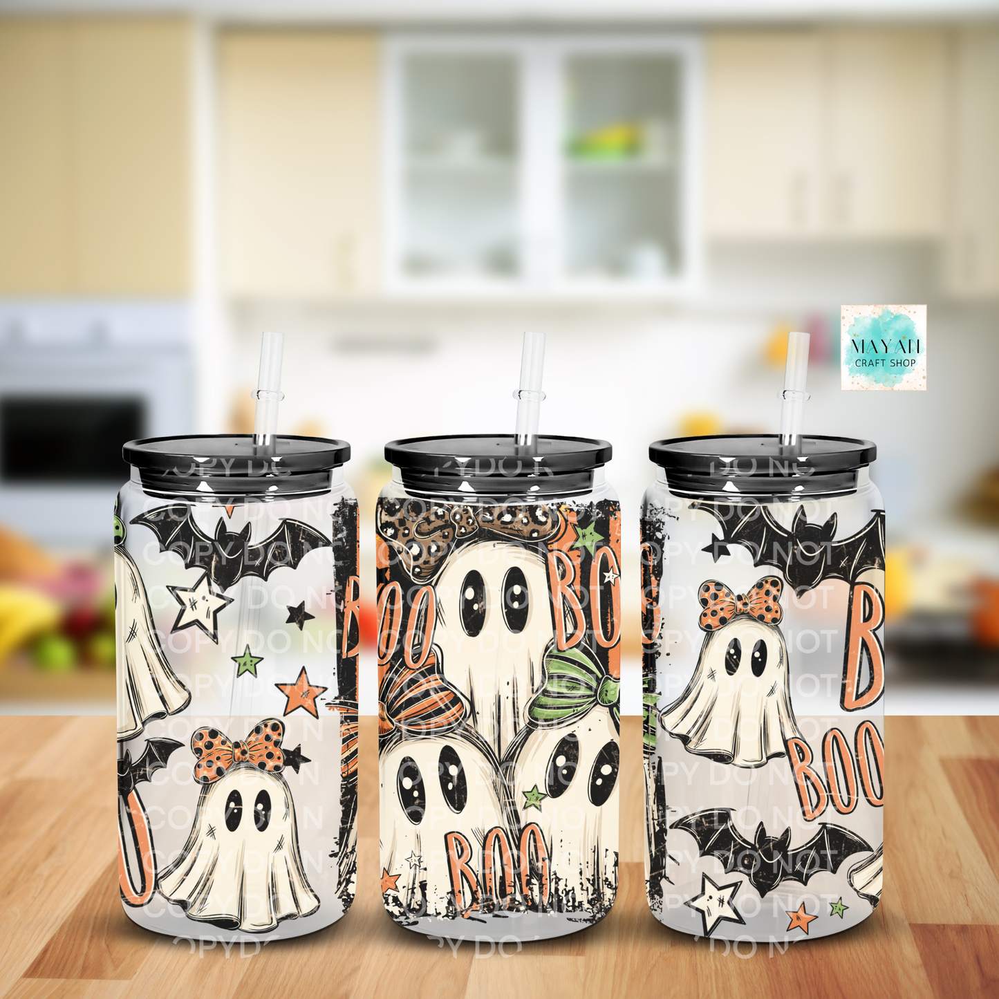 Three Halloween-themed stainless steel tumblers with lids and straws, decorated with cartoon ghosts, bats, stars, and the word 'BOO' in orange, shown on a wooden surface with a blurred kitchen background.
