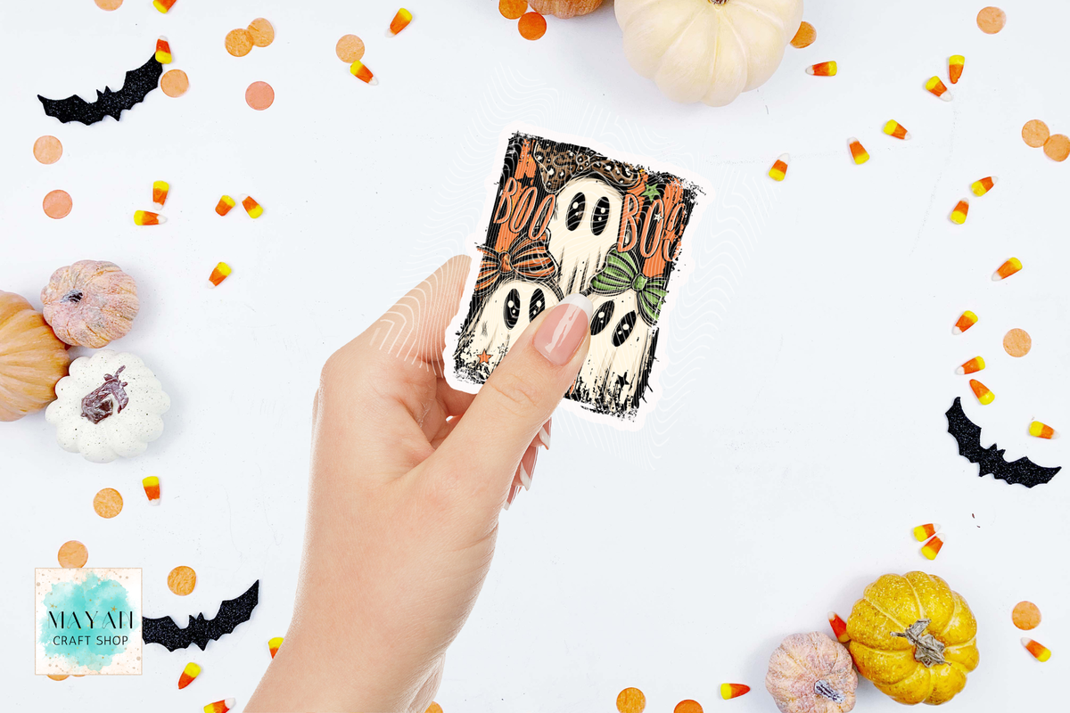A hand holds a square Halloween-themed sticker featuring three cartoon ghosts with 'BOO' text, surrounded by pumpkins, candy corn, and black bat cutouts on a white background. 