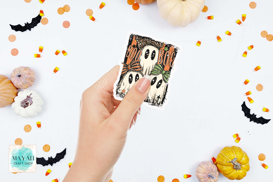 A hand holds a square Halloween-themed sticker featuring three cartoon ghosts with 'BOO' text, surrounded by pumpkins, candy corn, and black bat cutouts on a white background. 
