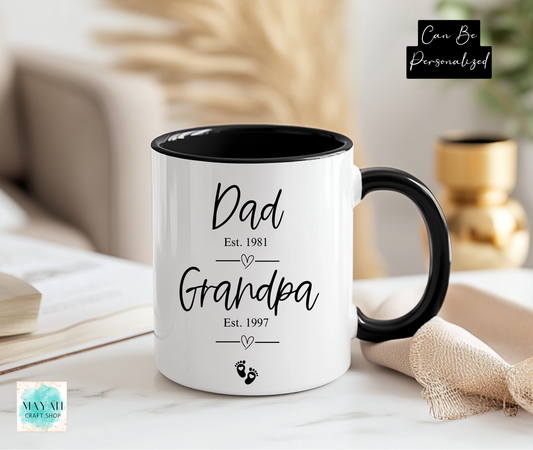 White mug with black interior and handle displaying 'Dad Est. 1981', 'Grandpa Est. 1997' text on a blurred home background.