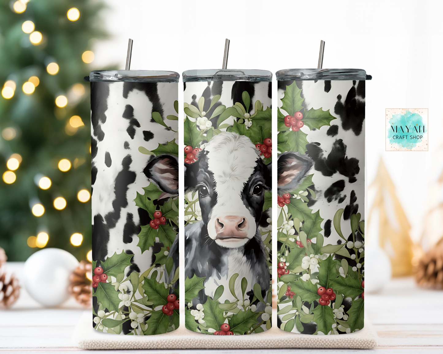 Three tumblers with cow designs and holly leaves on a decorative stand with a blurred Christmas tree background.