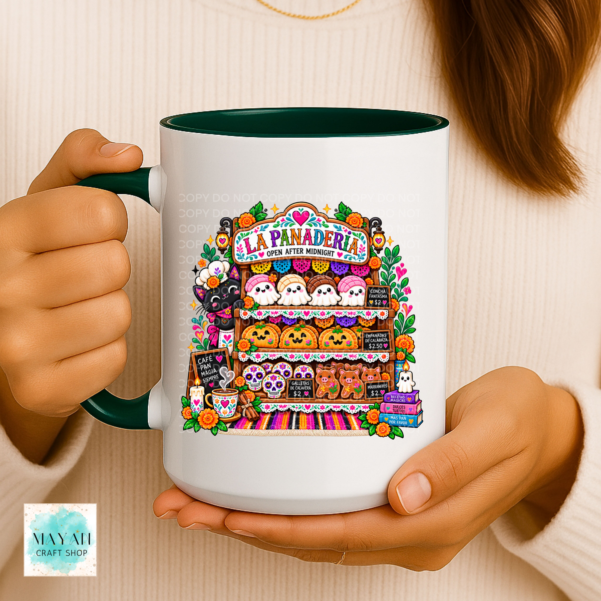 A person holds a white ceramic mug with a dark green handle and interior. The mug features a colorful Halloween-themed illustration of a candy store labeled 'La Panadería' with ghosts, pumpkins, and treats.