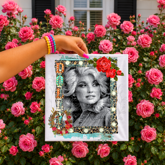 A hand wearing colorful bracelets holds a shirt in front of a dense background of pink roses. The canvas features a black-and-white portrait of a woman with a decorative multicolor frame and red roses.