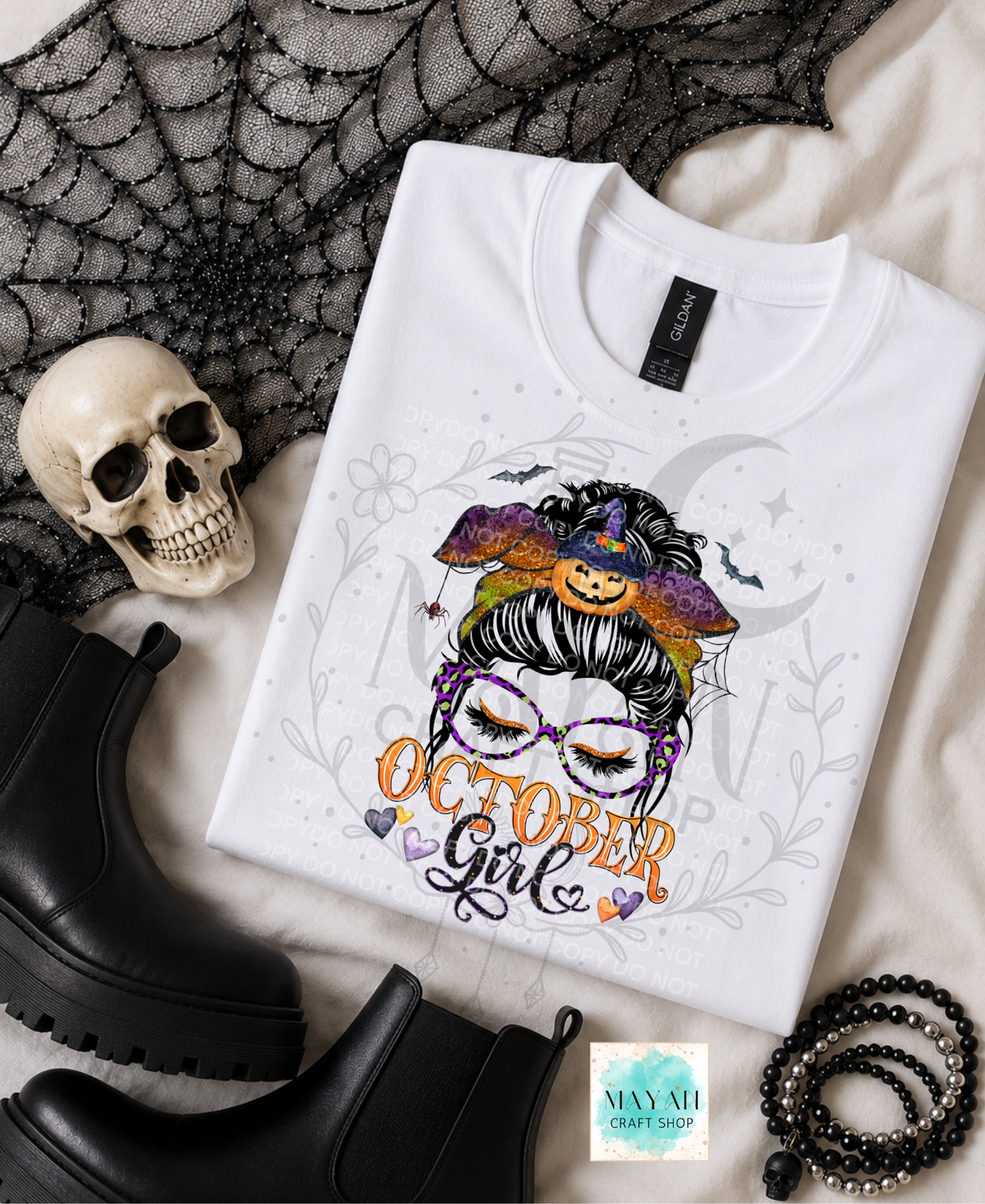 A white graphic T-shirt laid flat on a light fabric surface with Halloween-themed props, including a skull, black boots, a spiderweb print, and beads. The shirt features a stylized illustrated woman with glasses, a witch hat with a pumpkin, bats, and the words 'October Girl.