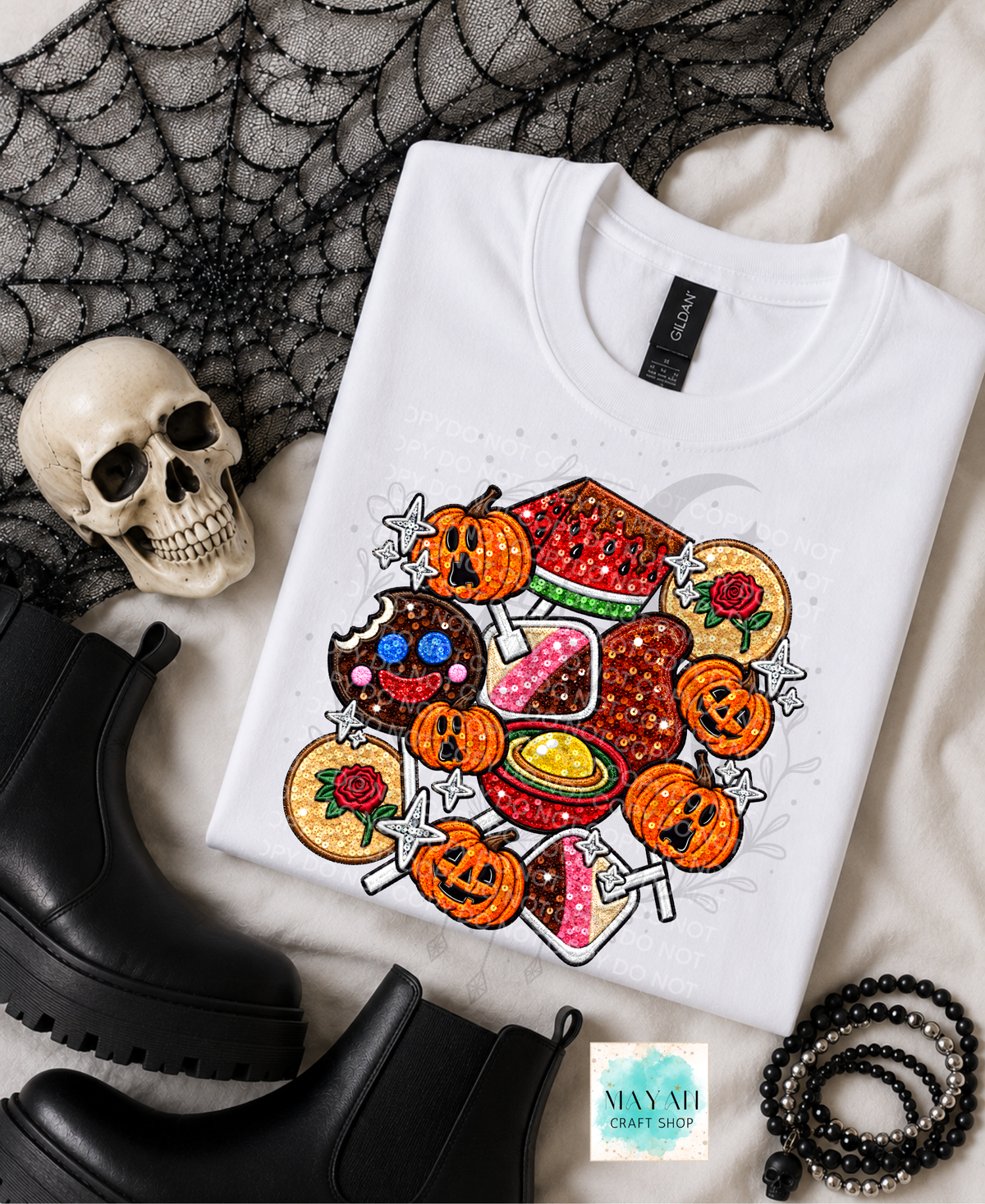 A white short-sleeve T-shirt laid flat on a light fabric surface with Halloween-themed props, including a skull, black boots, a spiderweb print, and beads. The shirt features a colorful graphic of mexican candy and other treats.