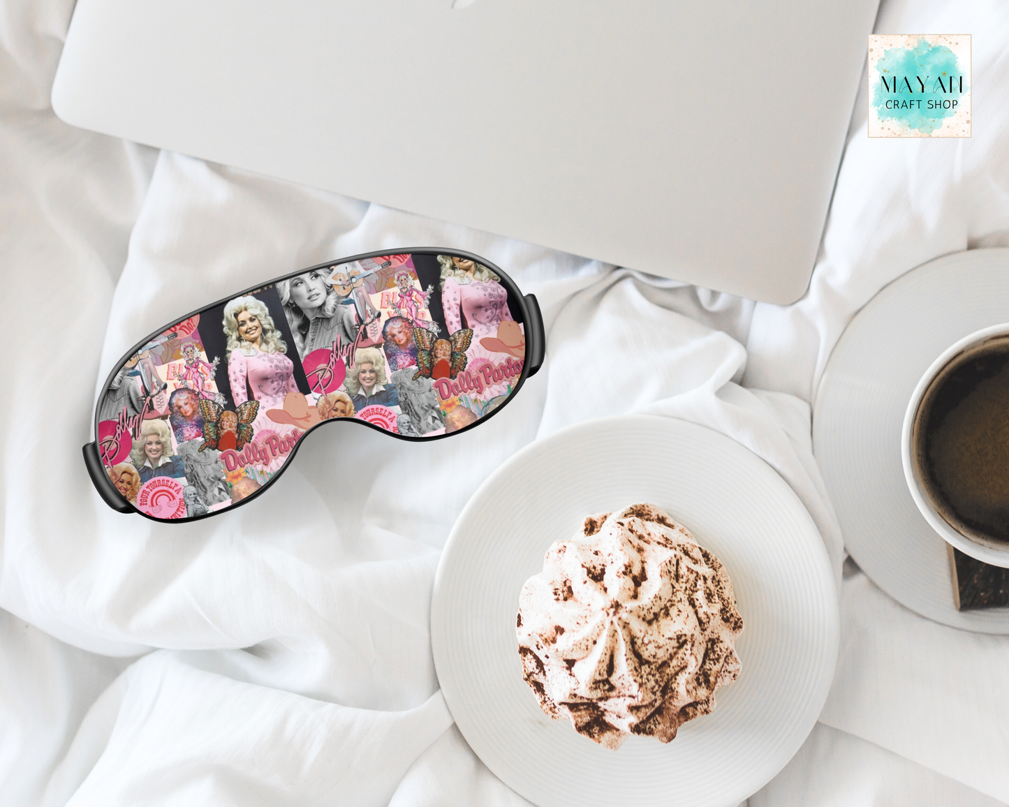 A sleep mask with a colorful collage print of vintage women in pink and floral patterns, styled on white bedding alongside a laptop, a cup of coffee, and a plate with a meringue dessert.