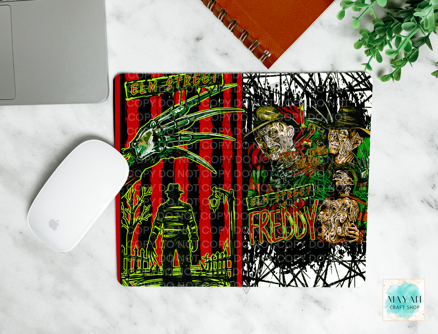 A mouse pad featuring a split horror design: a red-and-black striped man on the left and a black-and-white scribble-style portrait section on the right. The mat is shown on a white marble surface with part of a laptop, a white mouse, an orange notebook, and greenery visible around it.