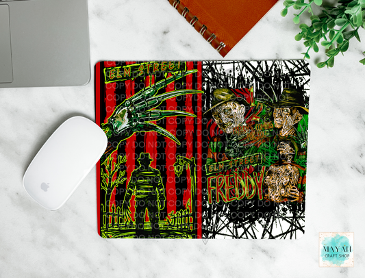 A mouse pad featuring a split horror design: a red-and-black striped man on the left and a black-and-white scribble-style portrait section on the right. The mat is shown on a white marble surface with part of a laptop, a white mouse, an orange notebook, and greenery visible around it.